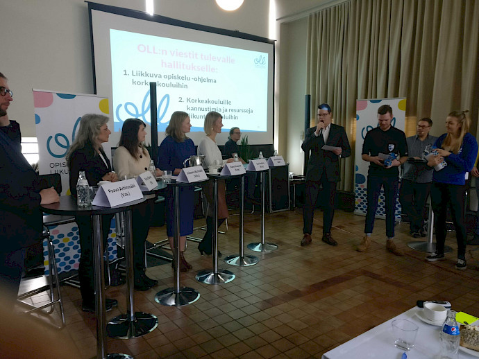 Panellists in Lasipalatsi&#39;s Palm Room at the start of the #liikkuvaopiskeluNYT parliamentary election panel organised by OLL in March 2019: Paavo Arhinmäki (Left Alliance), Ritva Elomaa (Finns Party), Johanna Karimäki (Green League), Hanna Kosonen (Centre Party), Sari Multala (National Coalition Party) and Joona Räsänen (Social Democratic Party of Finland). Talking with the panellists is Joonas Suikula, OLL’s President for 2019. Standing next to Joonas are Vice Presidents Harri Nurmi and Tiina Anttonen. Behind them are the presenters of the panel, OLL’s Special Advisor for University Sport Jussi Ansala and OLL’s President for 2018 Soile Koski-Aho.